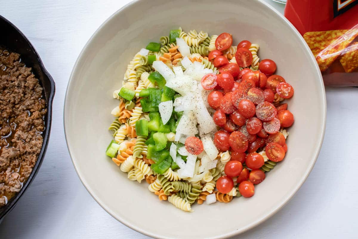 Overhead shot of cooked spiral pasta, chopped green bell pepper, chopped onion, and halved grape tomatoes.