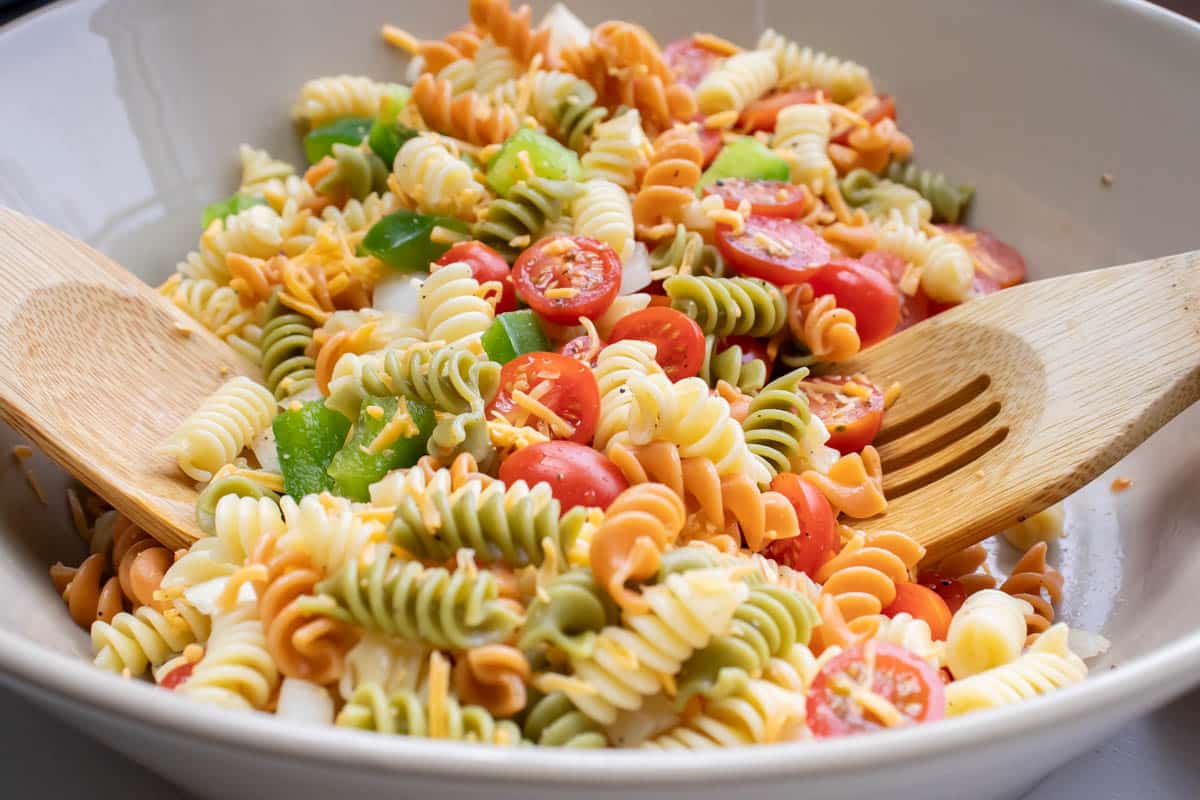 Multi-colored rotini noodles in a bowl with chopped tomatoes and bell peppers and wooden salad spoons.