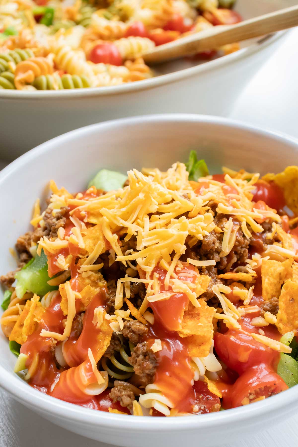Bowl of seasoned ground beef, spiral pasta, lettuce, halved tomatoes, nacho cheese Doritos, and French salad dressing with shredded cheddar cheese on top.