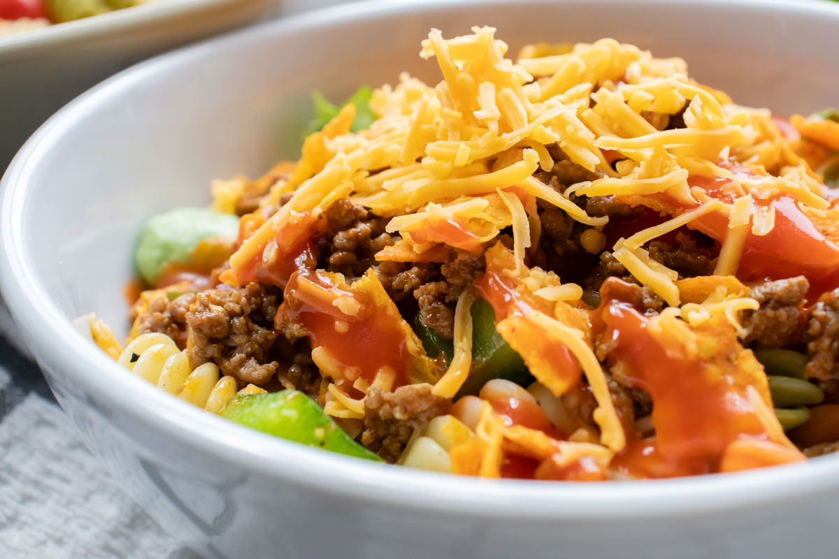 Bowl of seasoned ground beef, spiral pasta, lettuce, halved tomatoes, nacho cheese Doritos, and French salad dressing with shredded cheddar cheese on top.