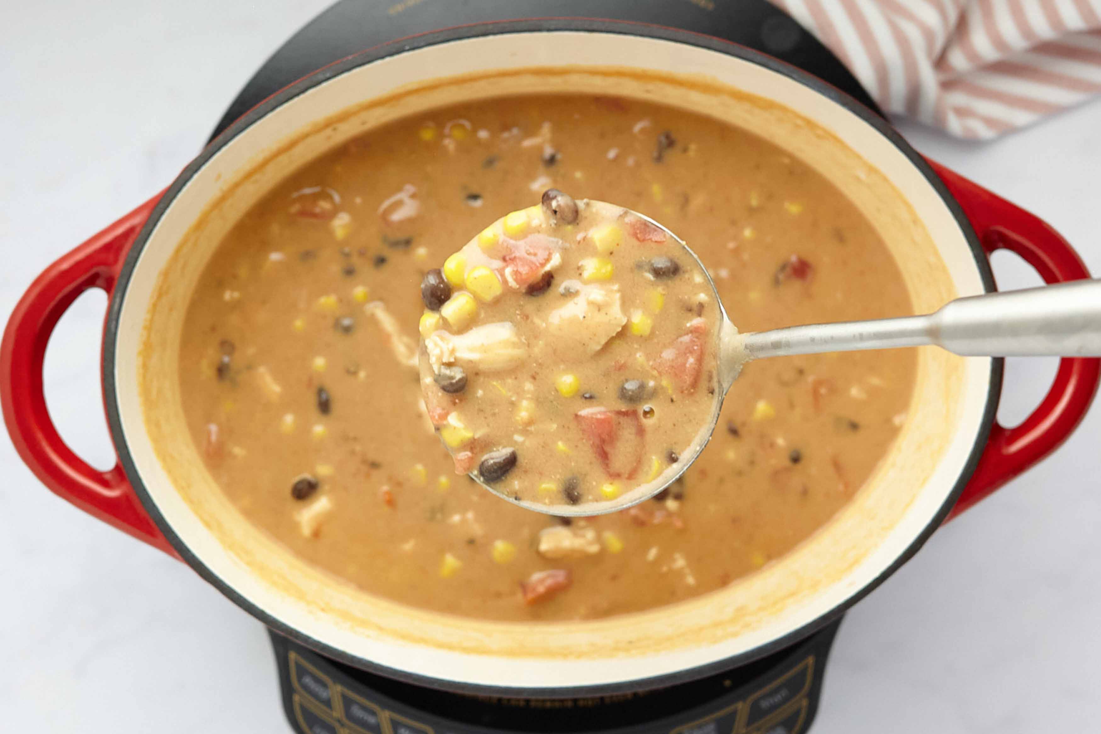 A ladleful of creamy soup showing chicken, diced tomatoes, black beans, and corn held above a Dutch oven full of the soup.