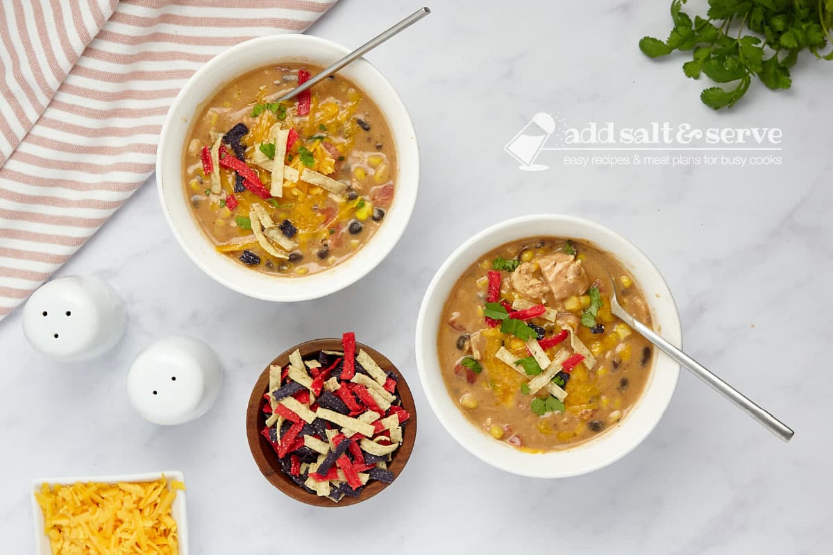 Two bowls of chicken enchilada soup garnished with shredded cheddar cheese, cilantro, and tortilla strips.