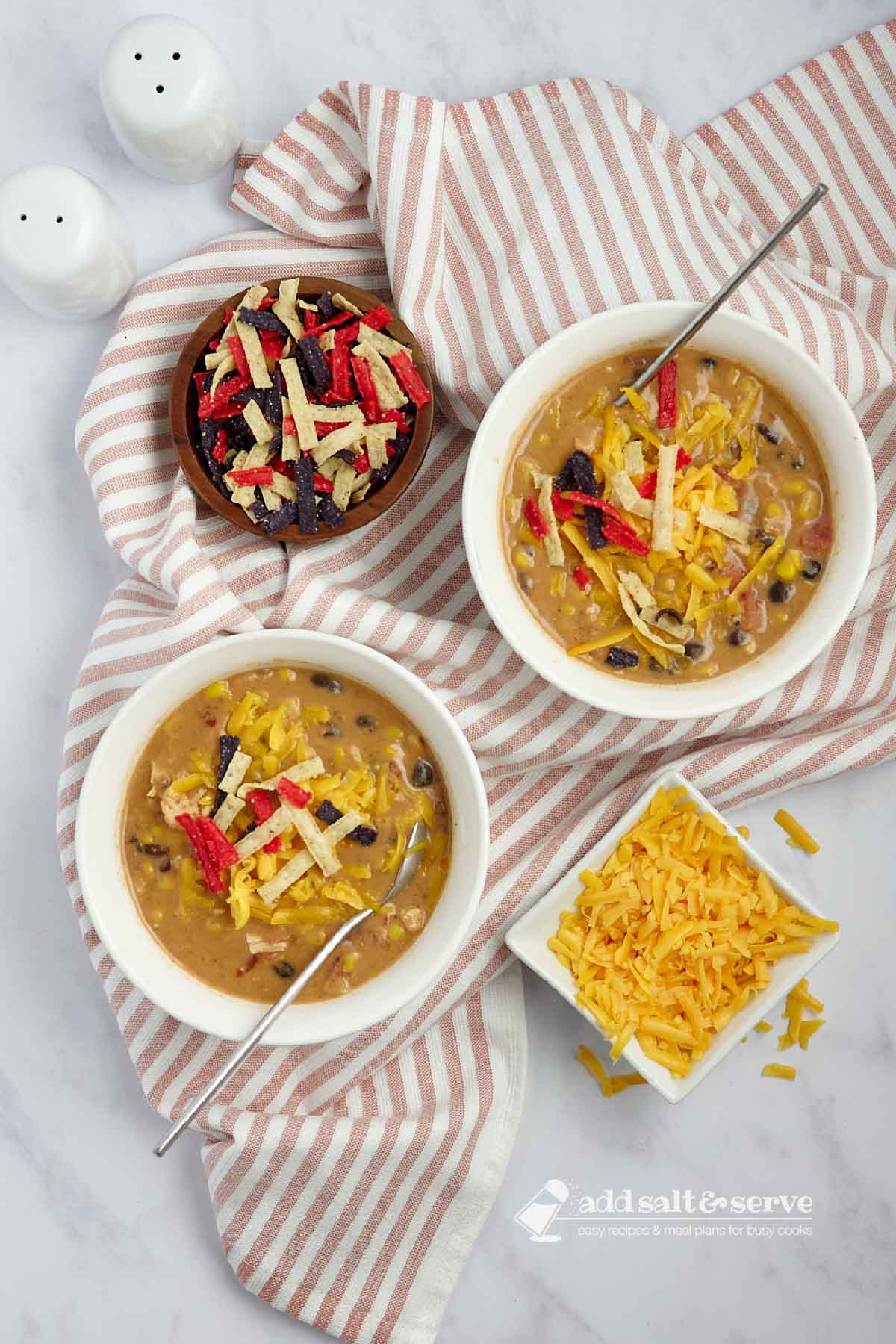 Two bowls of chicken enchilada soup garnished with shredded cheddar cheese, cilantro, and tortilla strips beside small bowls of shredded cheddar cheese and tortilla strips.