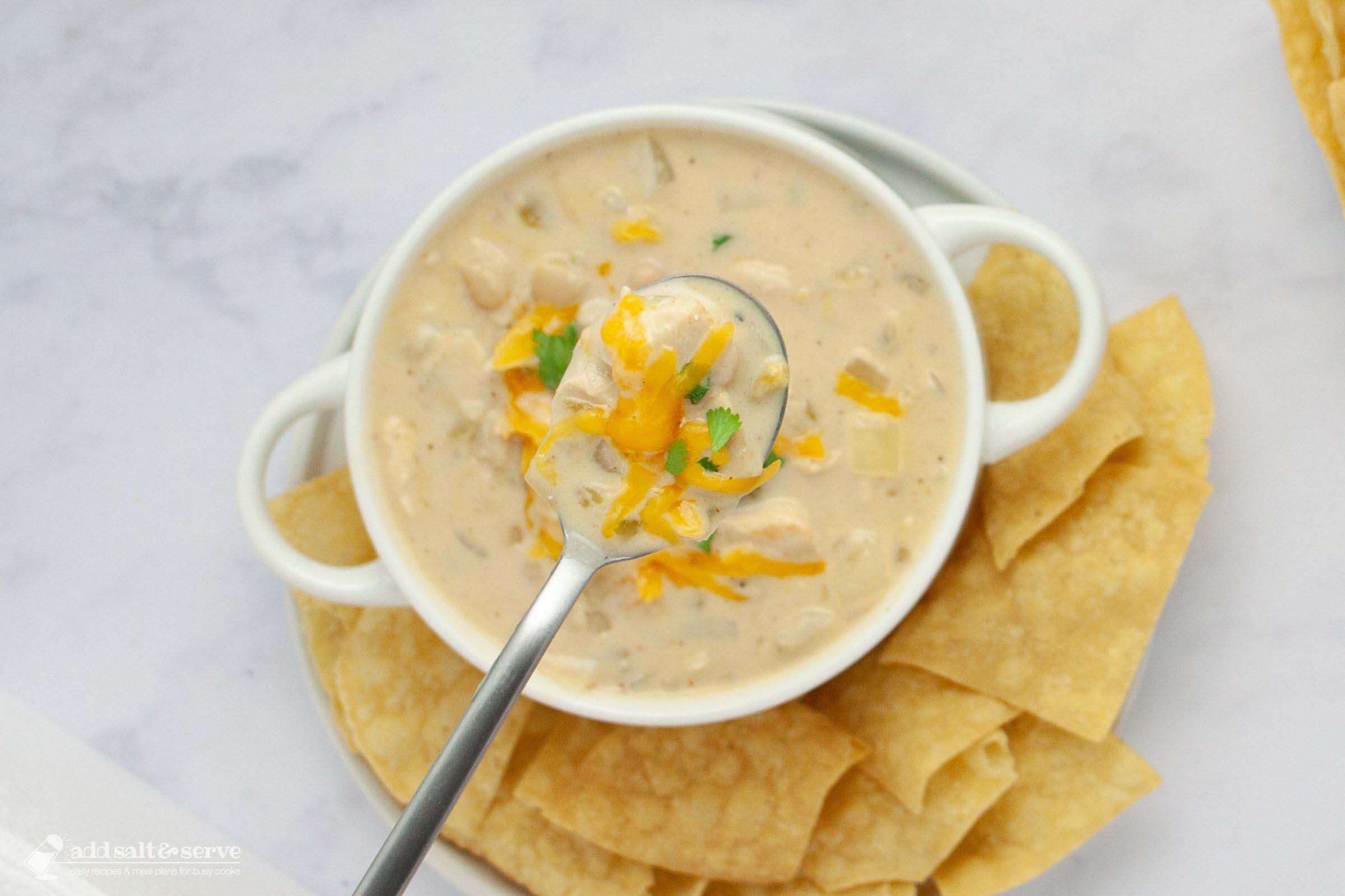 A bowl of creamy white chicken chili garnished with cheddar cheese and cilantro and served with chips with a spoonful held above the bowl toward the camera.