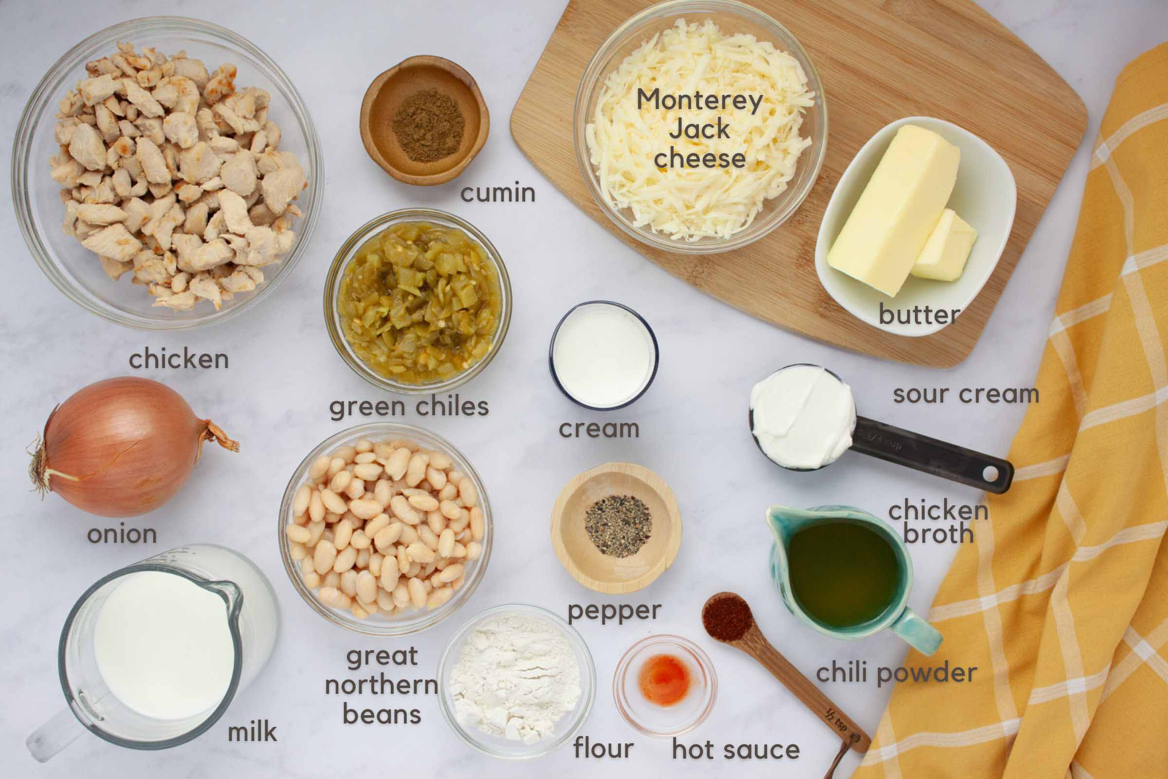 Labeled image of ingredients for creamy white chicken chili: chicken, onion, milk, cream, chicken broth, great northern beans, green chiles, flour, butter,Monterey Jack Cheese. cumin, pepper, chili powder, hot sauce, and sour cream.