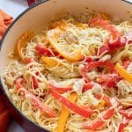 Pasta with chicken, sliced onion, and strips of red and yellow bell peppers cooked in a white sauce in a skillet.