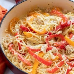 Pasta with chicken, sliced onion, and strips of red and yellow bell peppers cooked in a white sauce in a skillet.