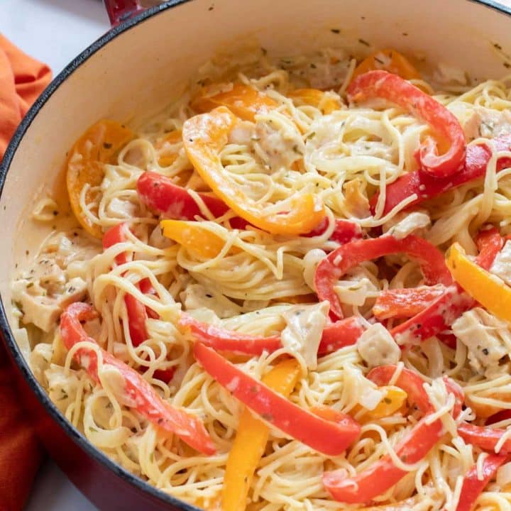 Pasta with chicken, sliced onion, and strips of red and yellow bell peppers cooked in a white sauce in a skillet.