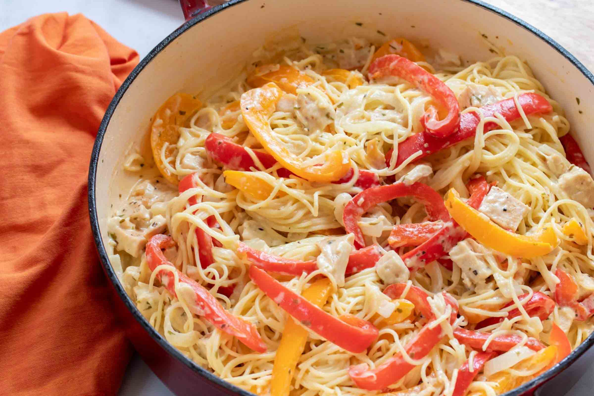 Pasta with chicken, sliced onion, and strips of red and yellow bell peppers cooked in a white sauce in a skillet.
