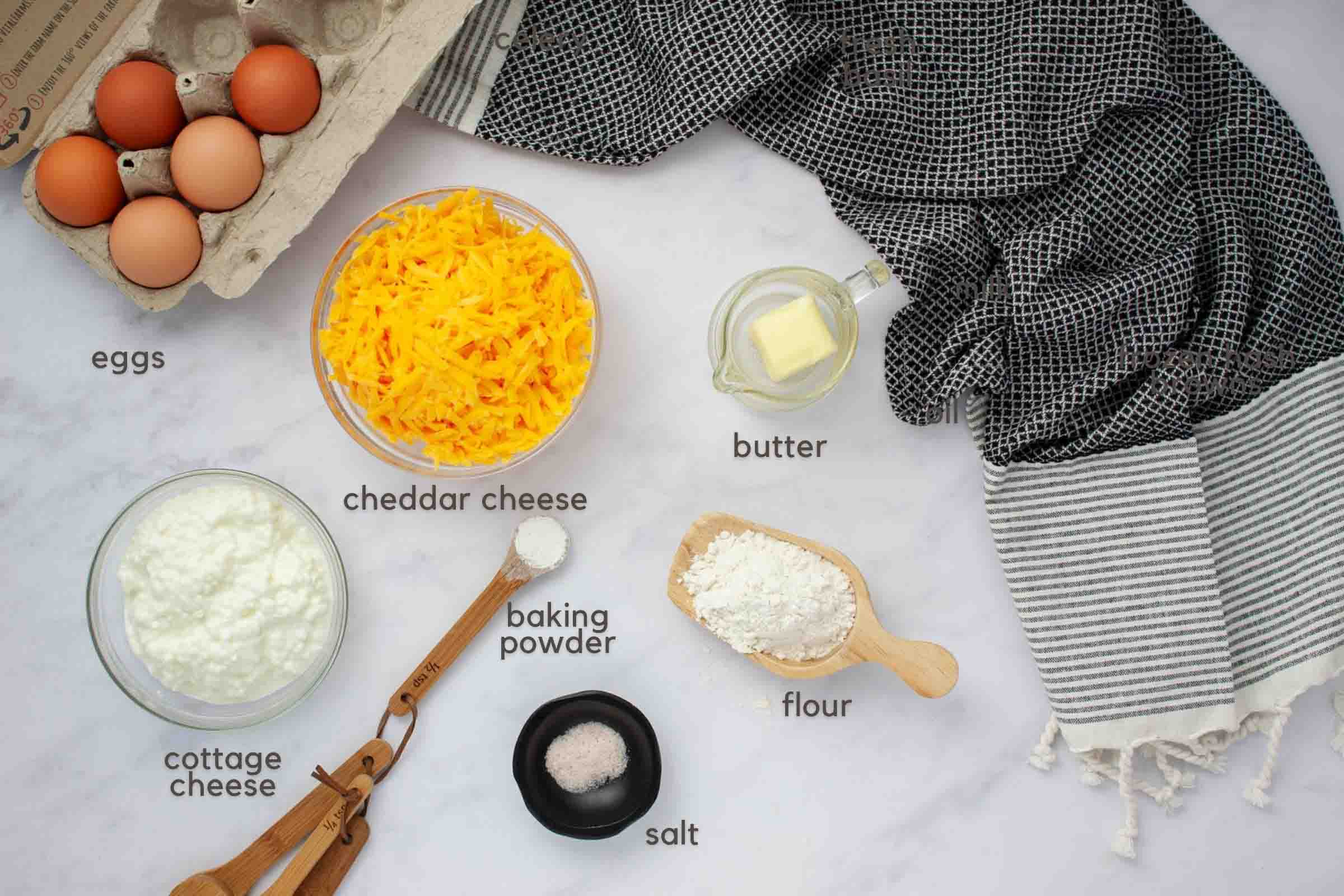 Ingredients to make quiche on a counter: eggs, shredded cheddar cheese, cottage cheese, baking powder, salt, flour, and butter.