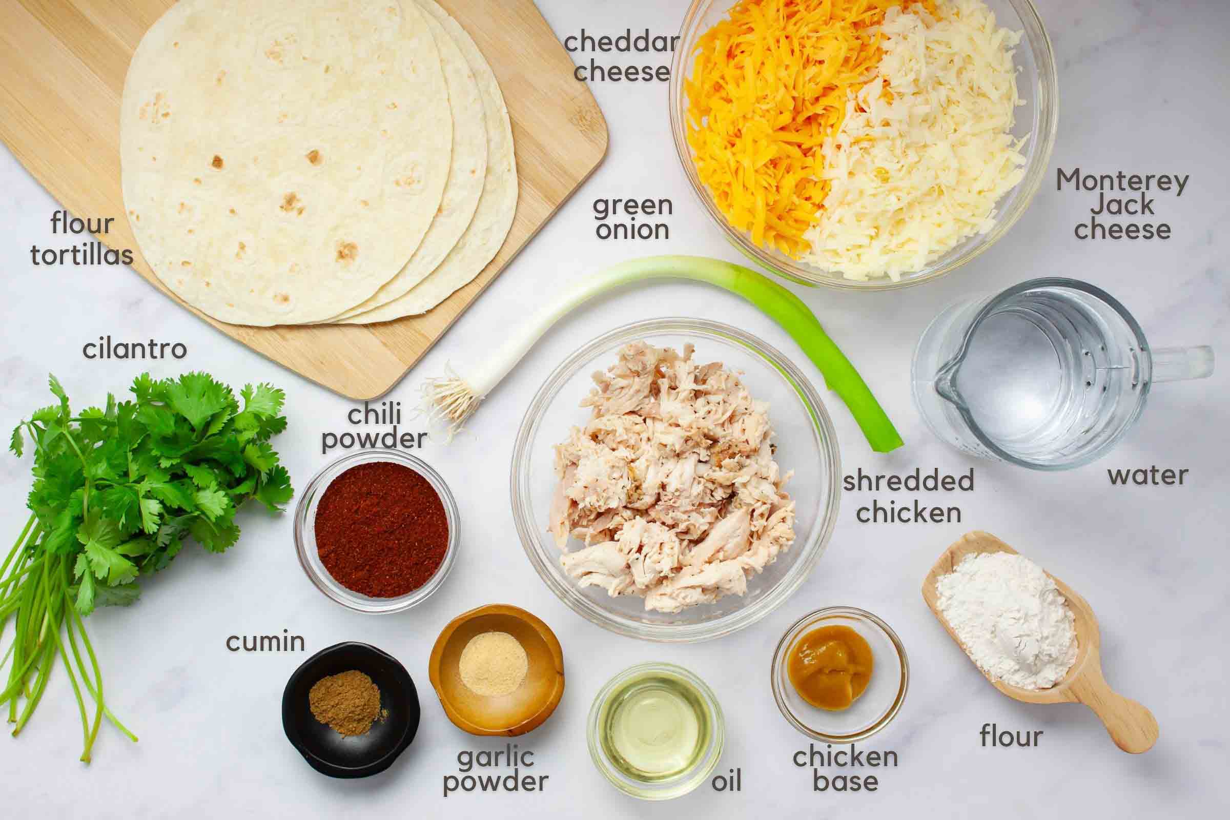 Ingredients on a counter: flour tortillas, shredded cheddar cheese, shredded Monterey Jack cheese, shredded rotisserie chicken, a green onion, fresh cilantro, chili powder, gorund cumin, garlic powder, oil, chicken base, all purpose flour, and water.