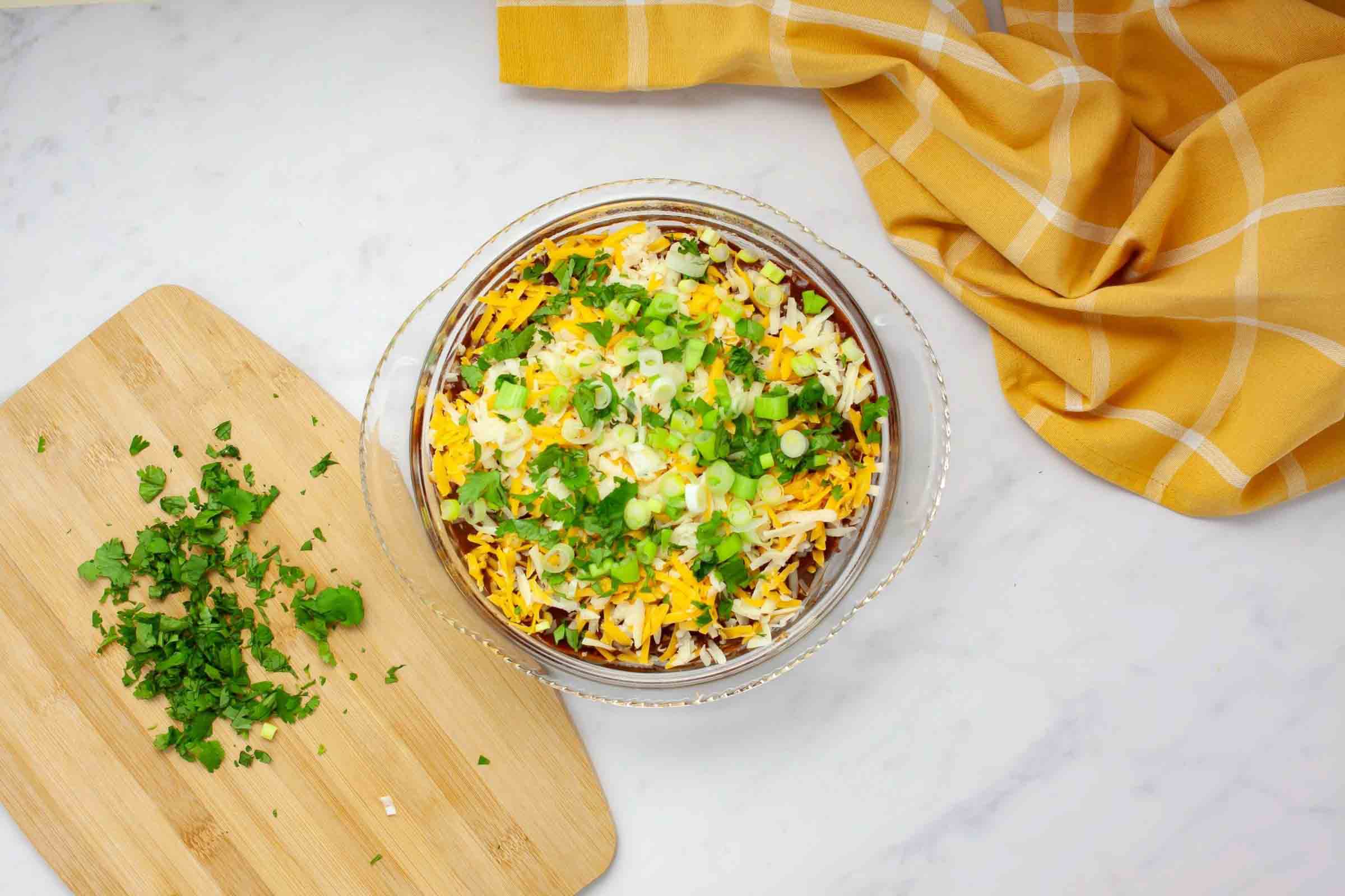 Overhead view of pie dish with layers of enchilada sauce, chicken, and cheese alternated with flour tortillas and topped with green onions and cilantro before baking.