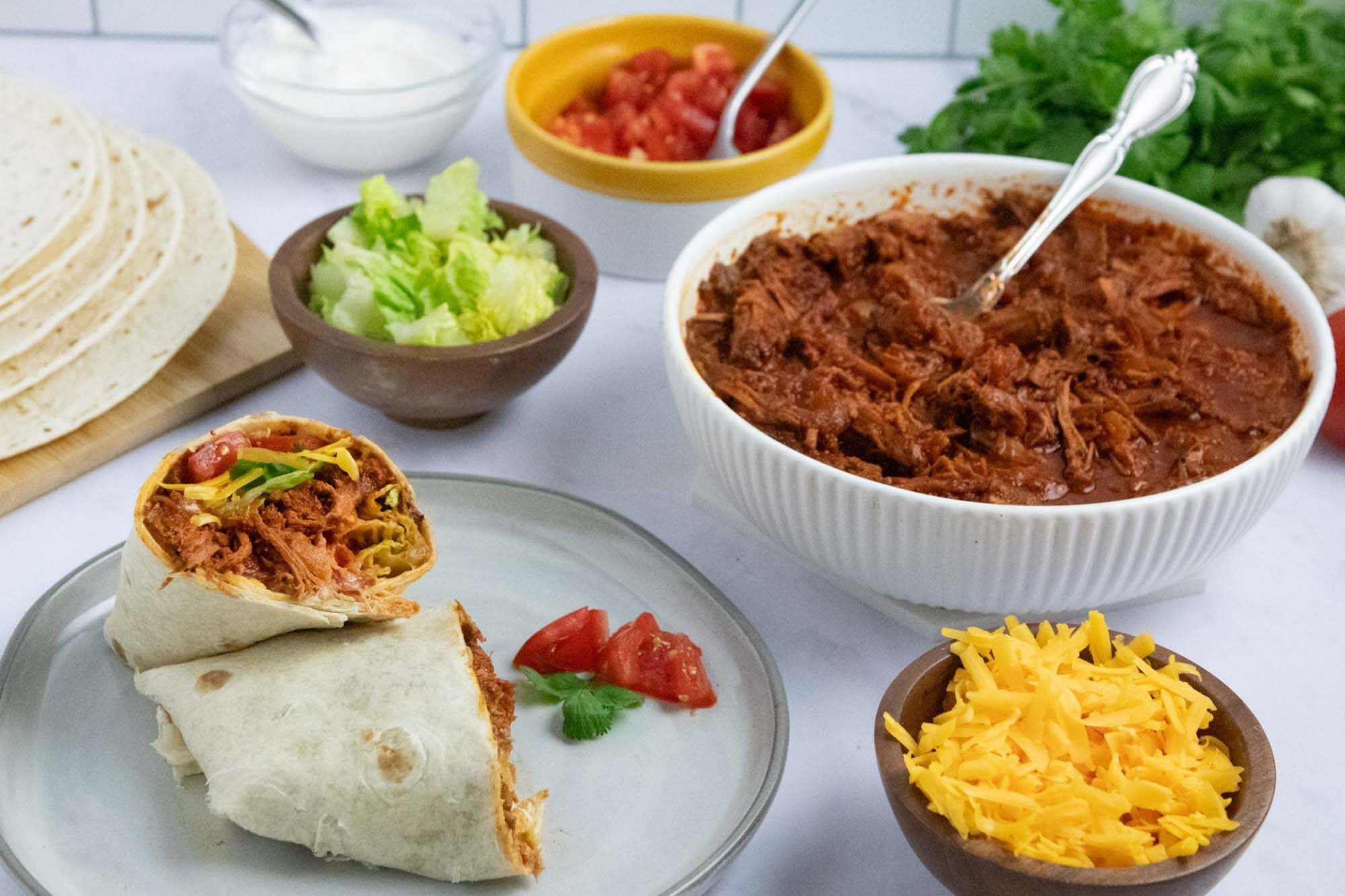 Overhead view of slow cooker beef burrito meat in a bowl surrounded by garnishes, tortillas, and a plate with an assembled burrito.