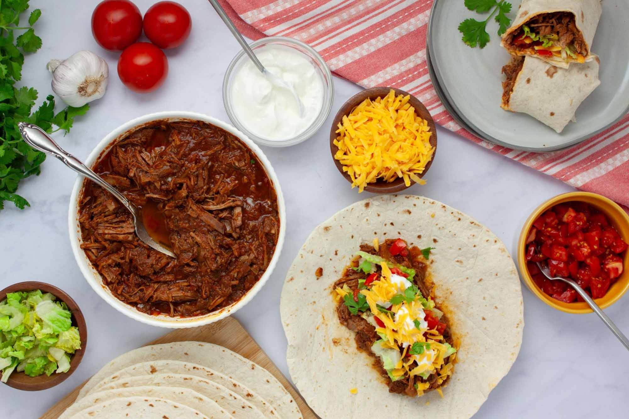 Overhead view of slow cooker beef burrito meat in a bowl surrounded by garnishes, tortillas, and a plate with a half-assembled burrito.