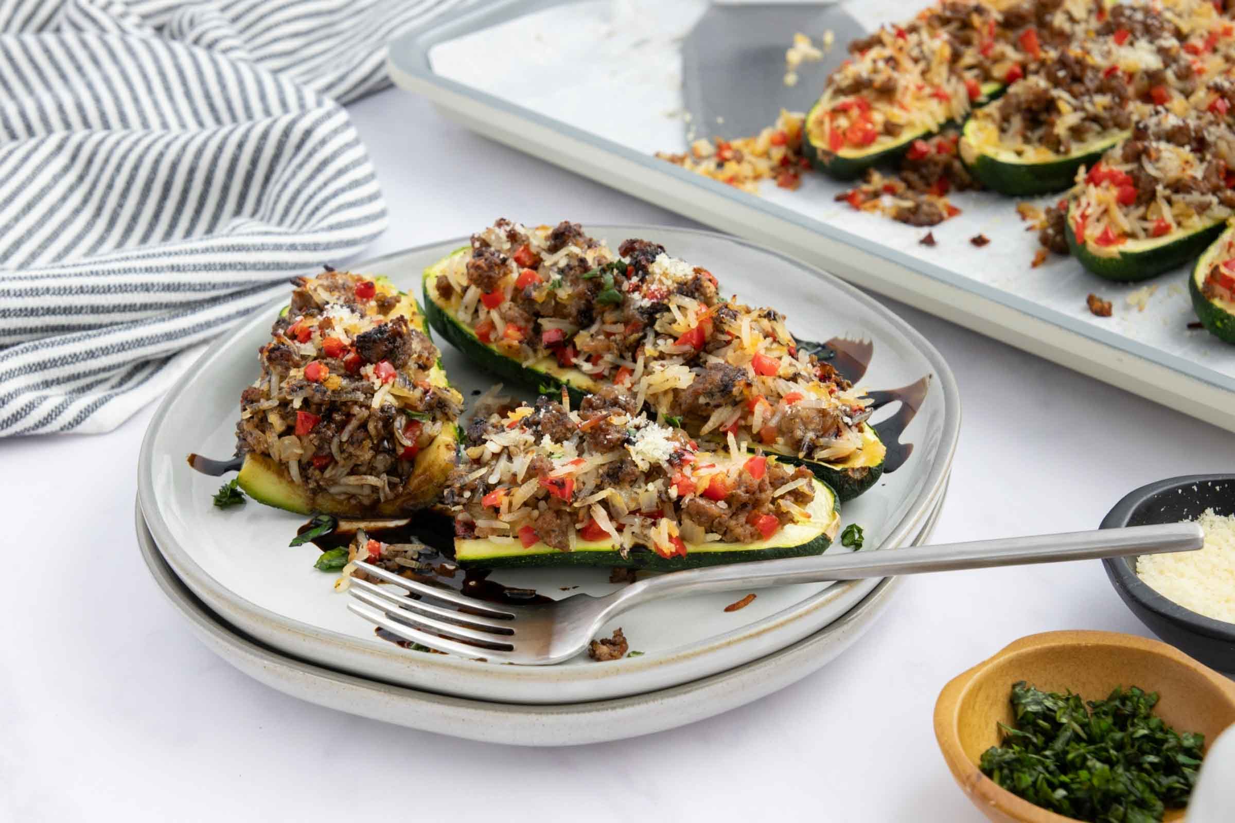 Baked zucchini stuffed with a rice, ground beef, and vegetable mixture drizzled with balsamic glaze on a plate next to a baking sheet lined with parchment paper with more zucchini.