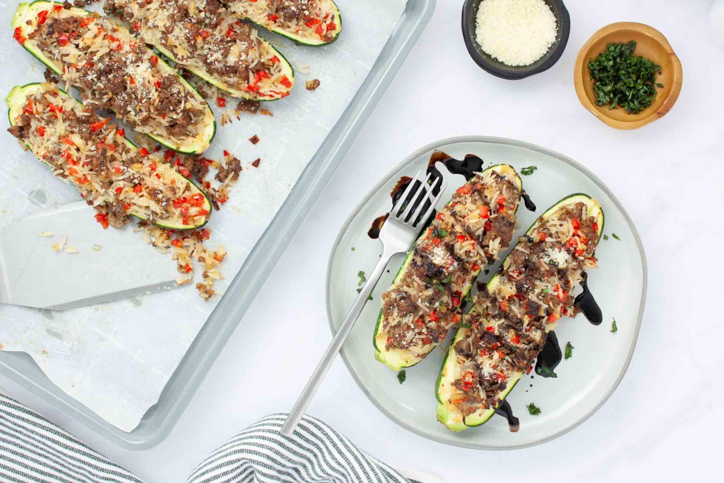 Baked zucchini stuffed with a rice, ground beef, and vegetable mixture drizzled with balsamic glaze on a plate next to a baking sheet lined with parchment paper with more zucchini.