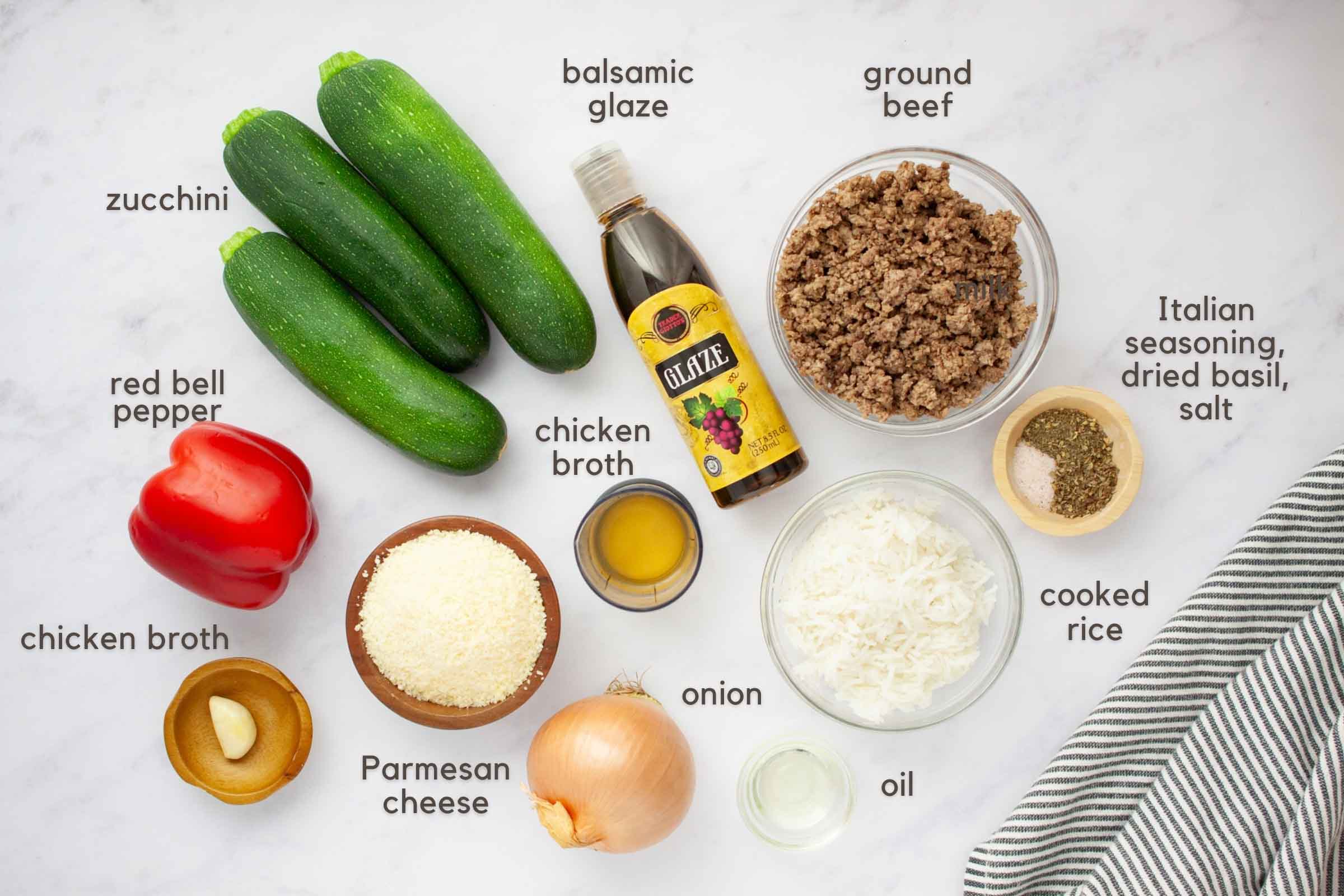 Ingredients on a counter: 3 zucchini, 1 red bell pepper, 1 onion, 1 clove garlic, bowl of Parmesan cheese, chicken broth, oil, cooked rice, cooked ground beef, Italian seasoning, dried basil, and salt.
