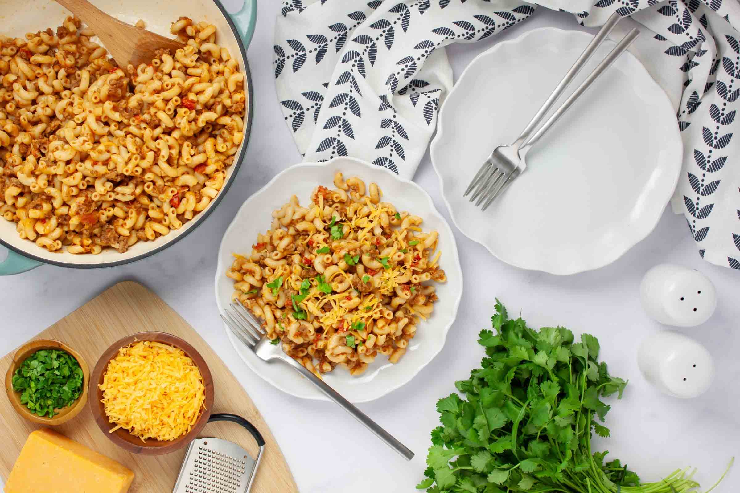 Pan of macaroni, beef, and tomatoes in a cheesy sauce beside a plate of the same garnished with cilantro and grated cheddar cheese.