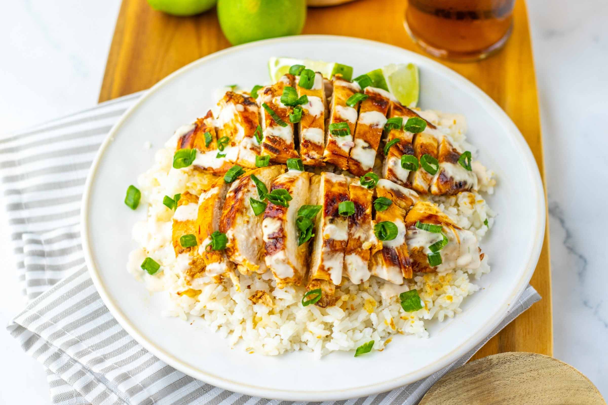 Chicken breast over rice drizzled with white sauce and garnished with green onions beside a glass of iced tea and two limes.