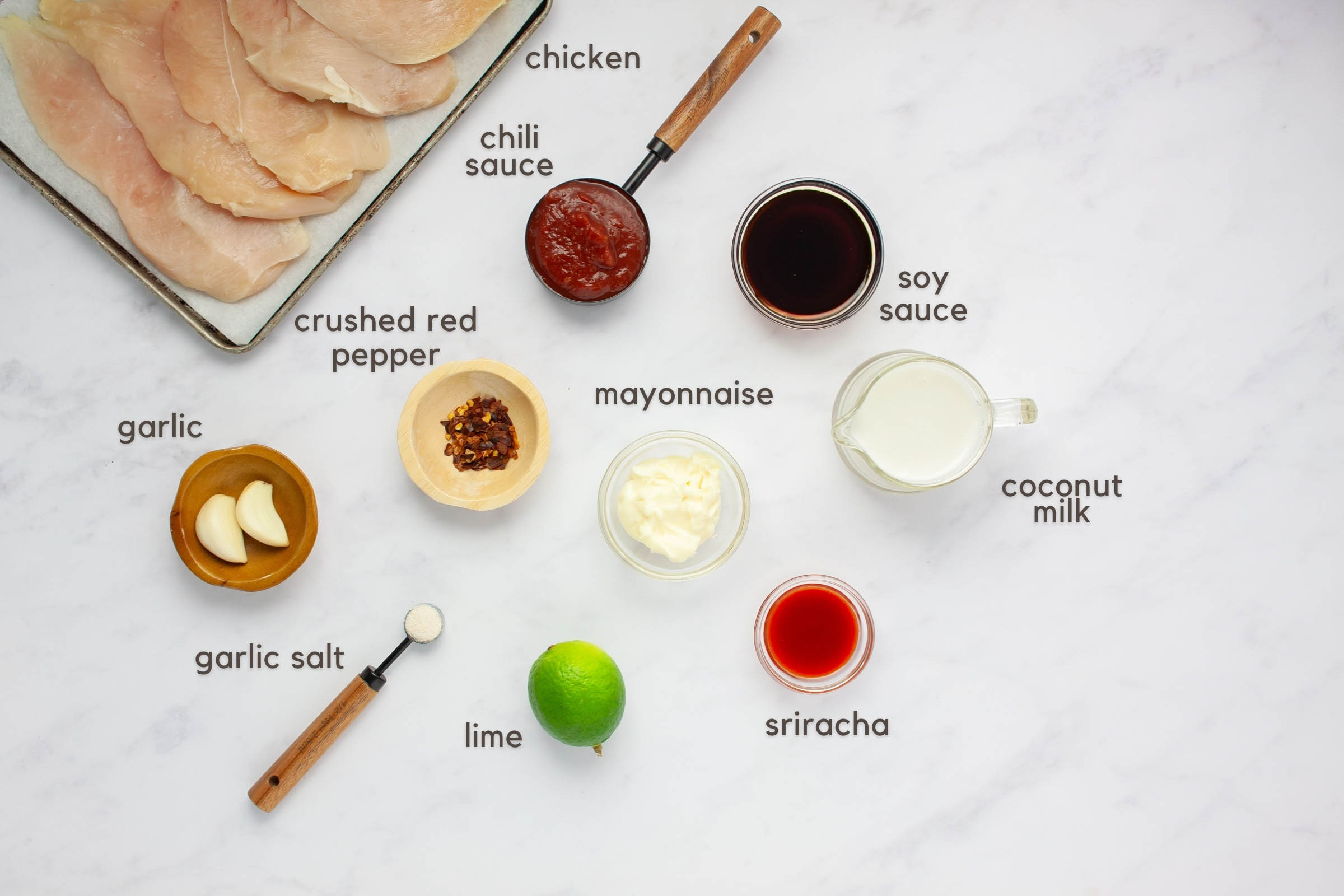 Labeled ingredients on a counter: boneless, skinless chicken breasts, chili sauce, soy sauce, crushed red pepper, garlic cloves, garlic salt, lime, mayonnaise, sriracha, and coconut milk.