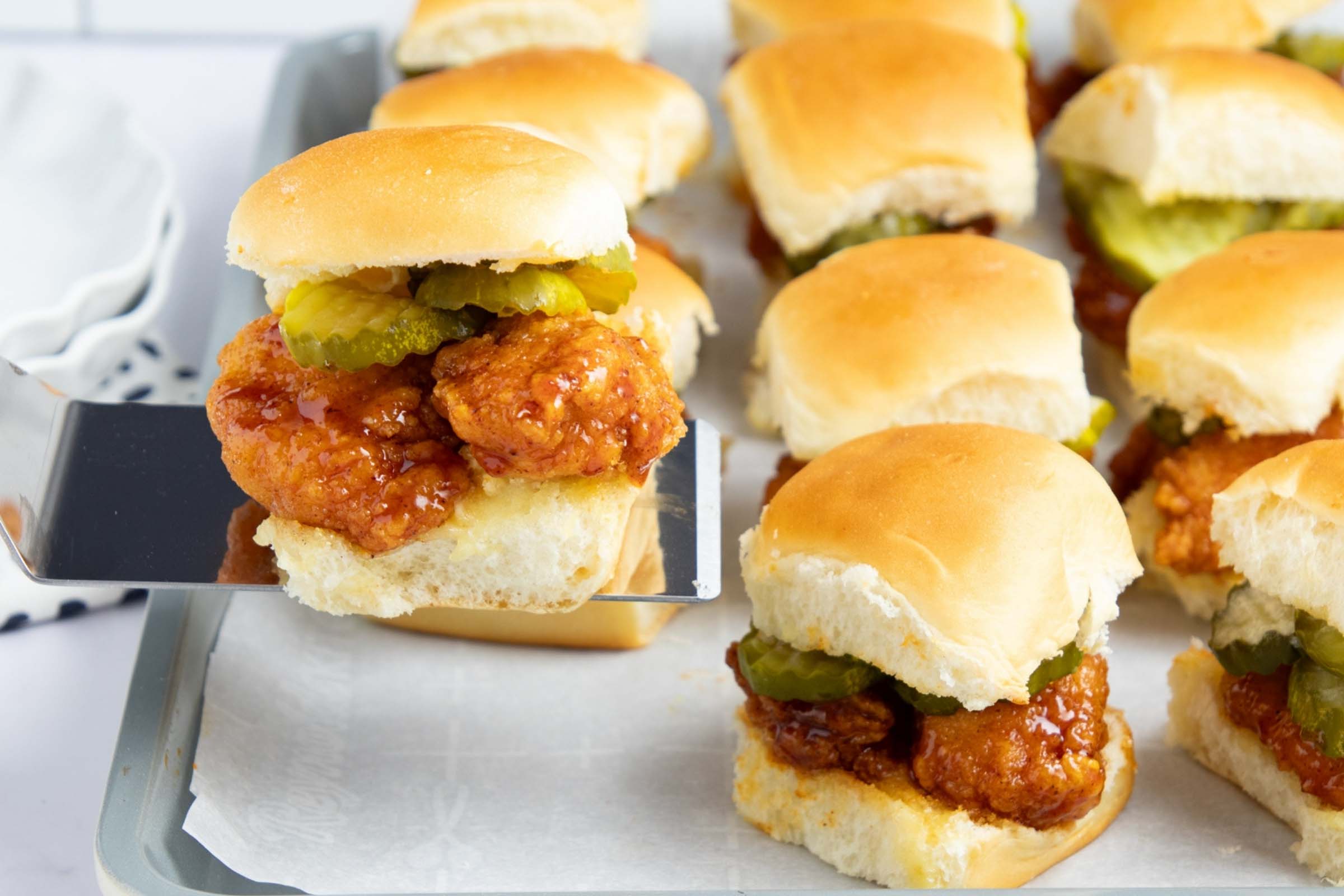 Hot honey chicken sliders with pickles on a baking sheet with one lifted on a spatula.