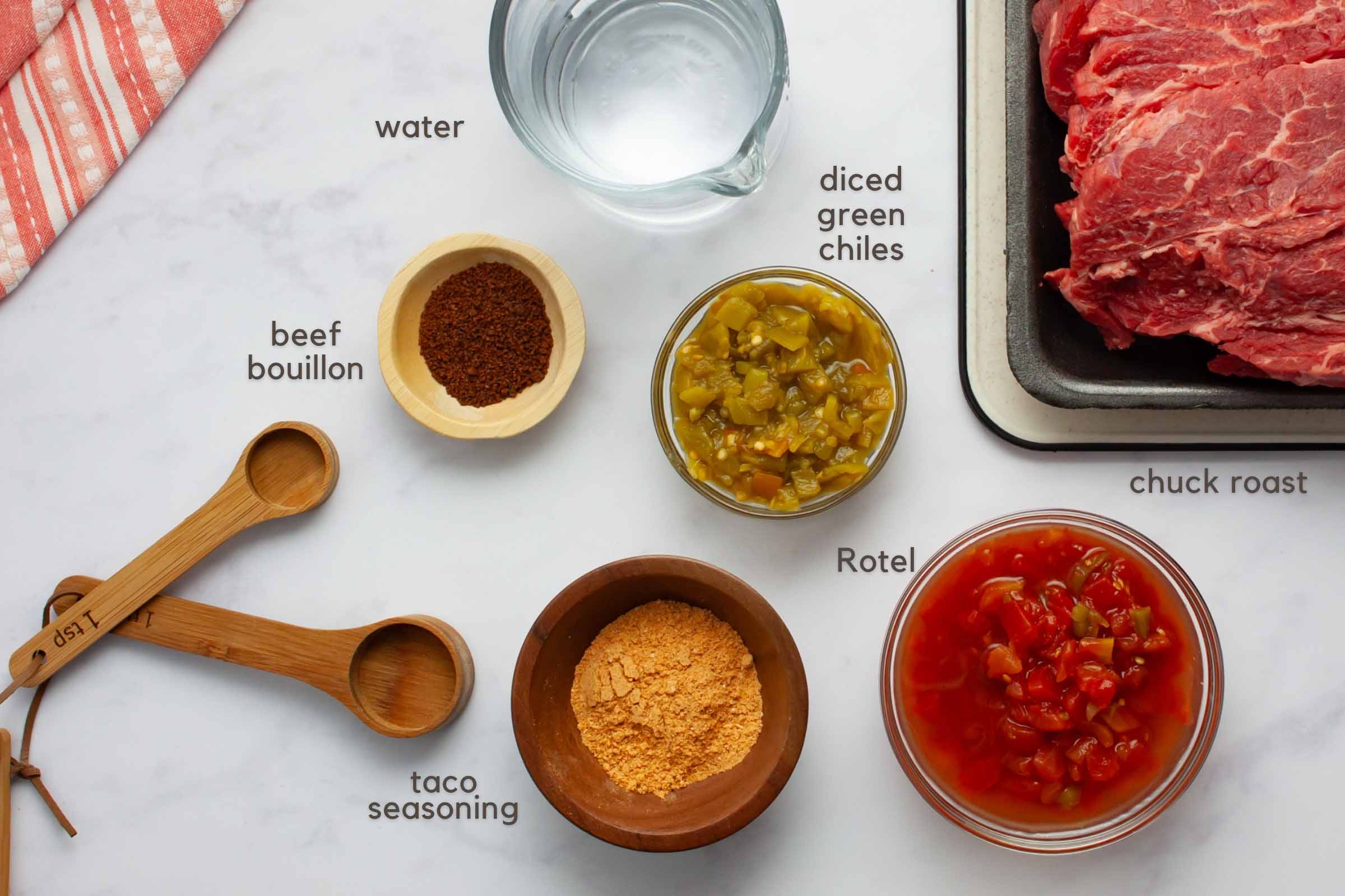 Ingredients on a counter: chuck roast, beef bouillon granules, water, diced green chiles, Rotel, and taco seasoning.