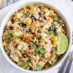 Bowl of chicken salad with corn, black beans, avocado, and red bell pepper in a creamy dressing garnished with a lime wedge and sprinkled with paprika.