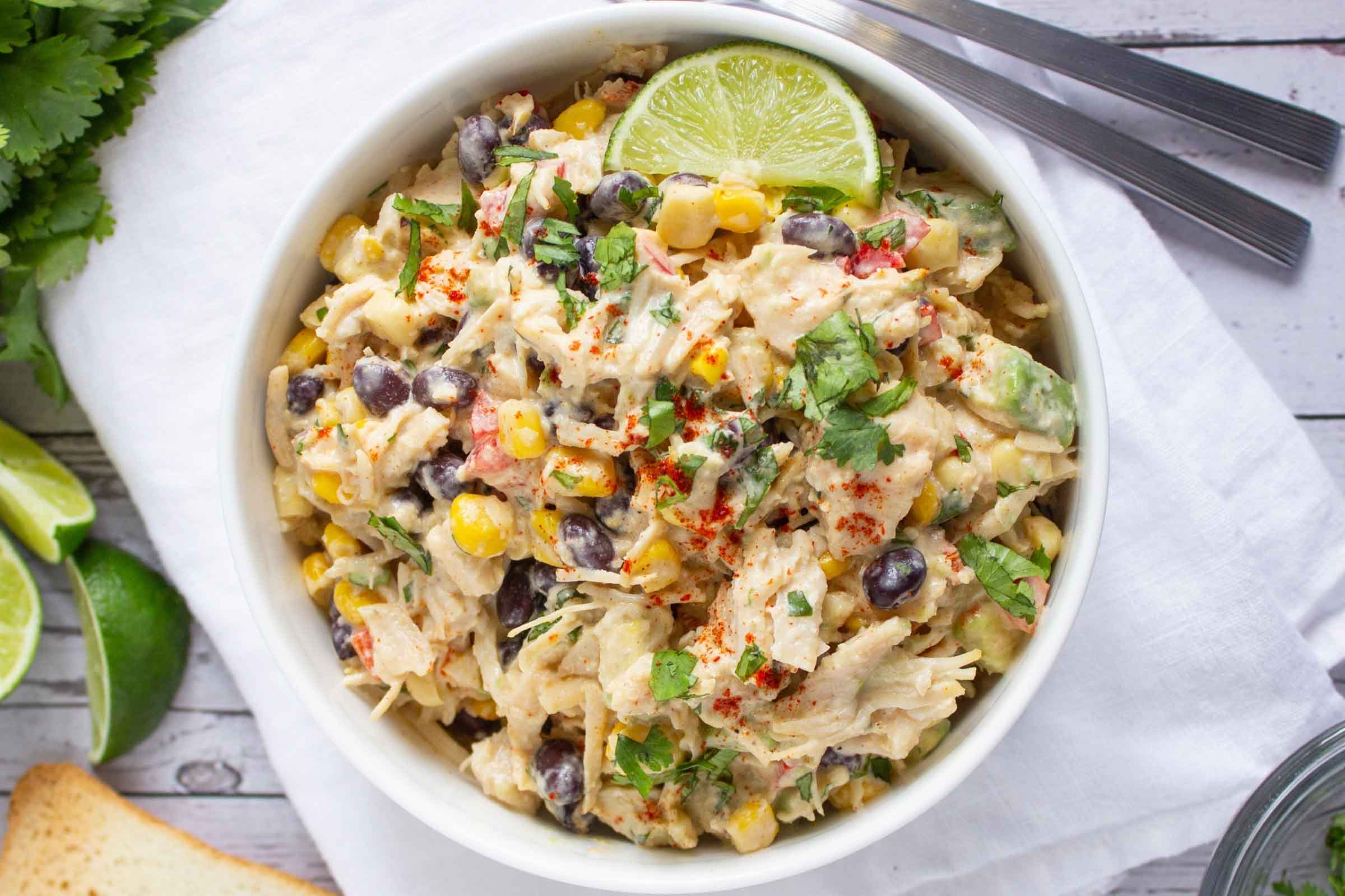 Bowl of chicken salad with corn, black beans, avocado, and red bell pepper in a creamy dressing garnished with a lime wedge and sprinkled with paprika.