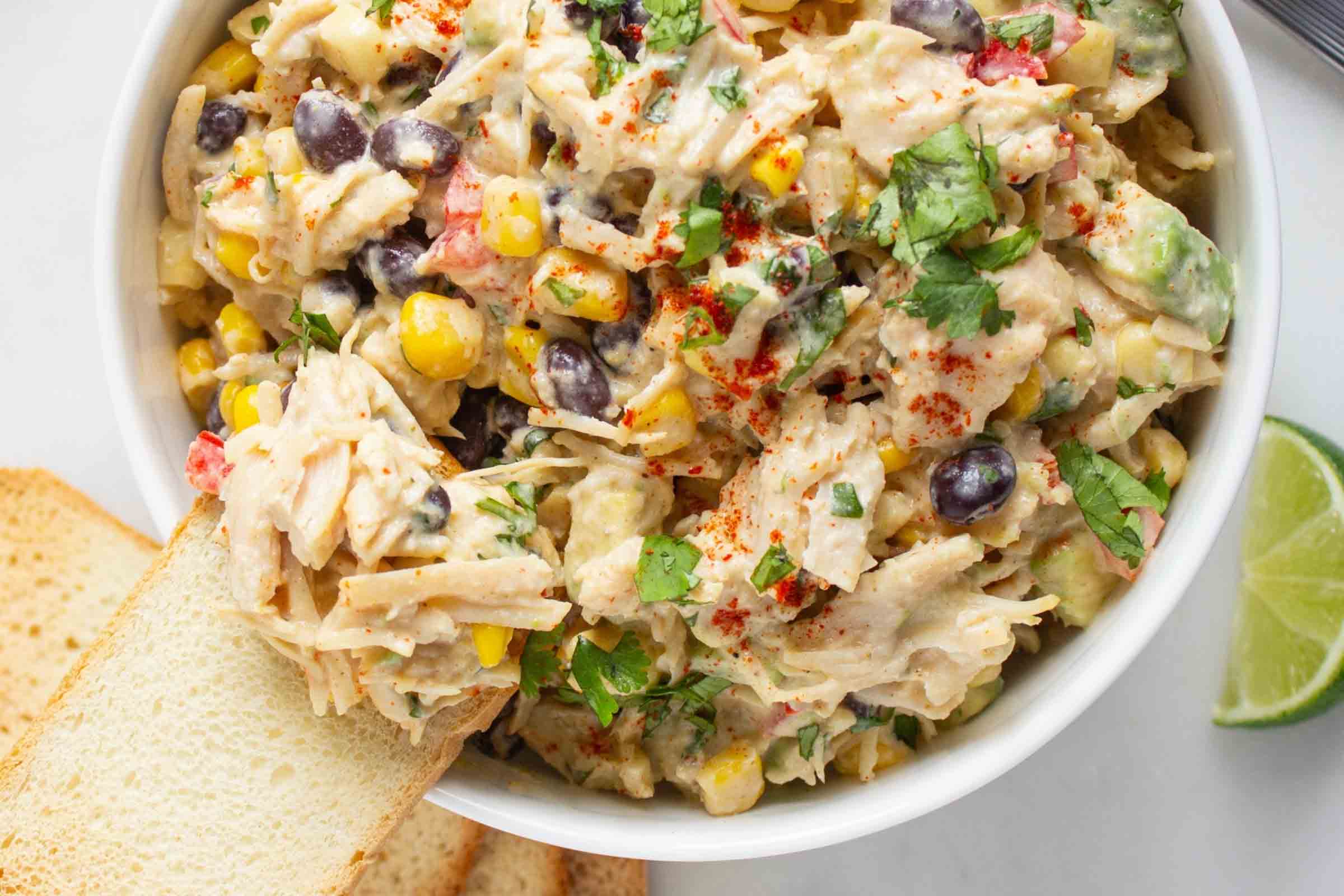 Bowl of chicken salad with corn, black beans, avocado, and red bell pepper in a creamy dressing sprinkled with paprika beside crostini and a lime wedge.