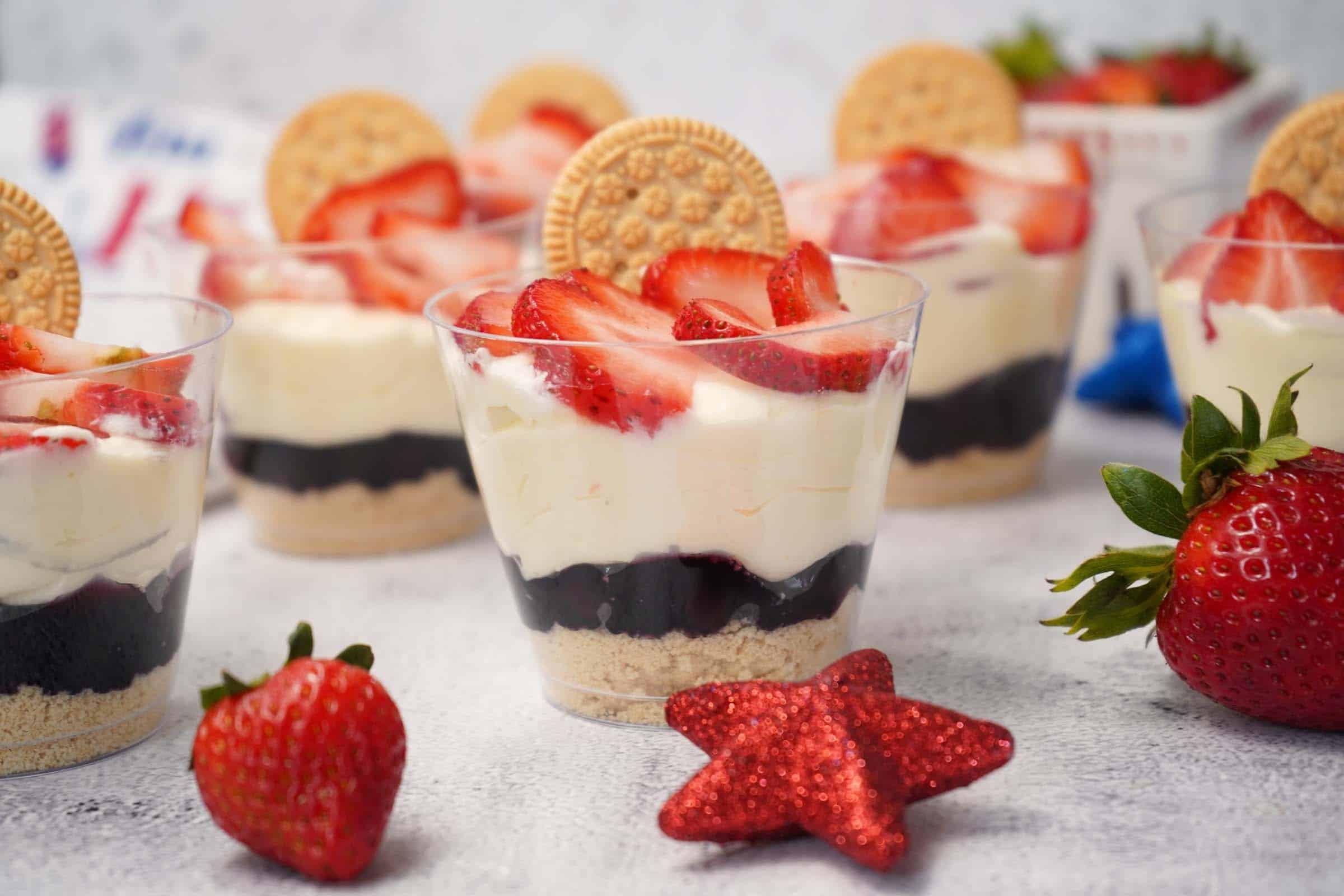 Red, white, and blue parfaits in plastic cups with layers of cookie crumbs, blueberry pie filling, pudding mixture, and topped with sliced strawberries and a sandwich cookie.