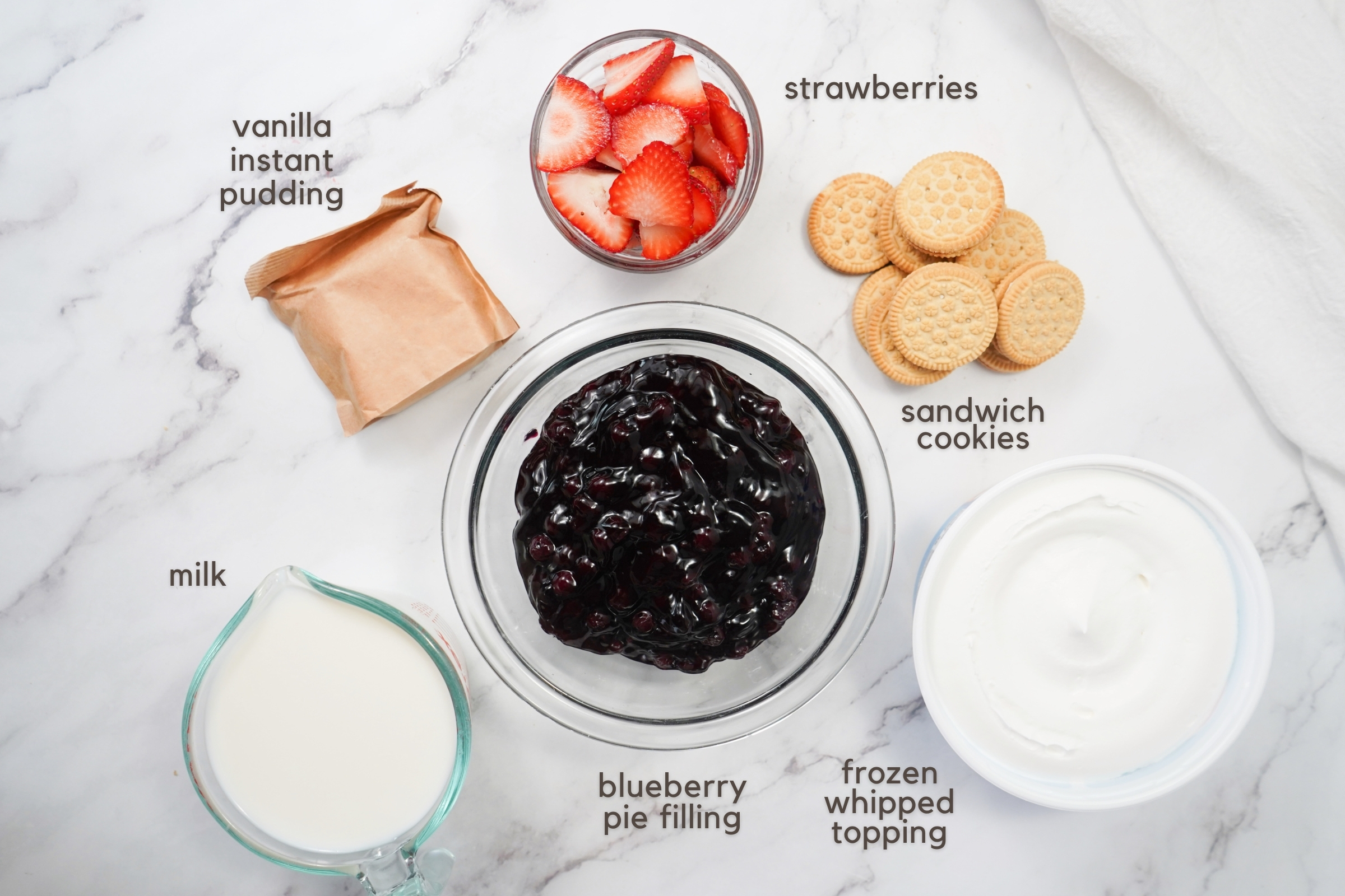 Labeled ingredients on a counter: Vanilla instant pudding mix, sliced strawberries, sandwich cookies, milk, blueberry pie filling, and frozen whipped topping.