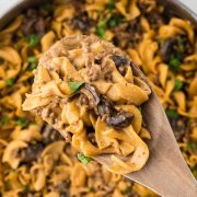 A pan with a finished recipe showing egg noodles, sliced mushrooms, and ground beef in a sauce garnished with chopped parsley with a serving lifted toward the camera on a wooden spoon.