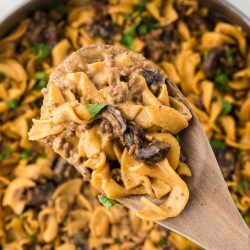 A pan with a finished recipe showing egg noodles, sliced mushrooms, and ground beef in a sauce garnished with chopped parsley with a serving lifted toward the camera on a wooden spoon.