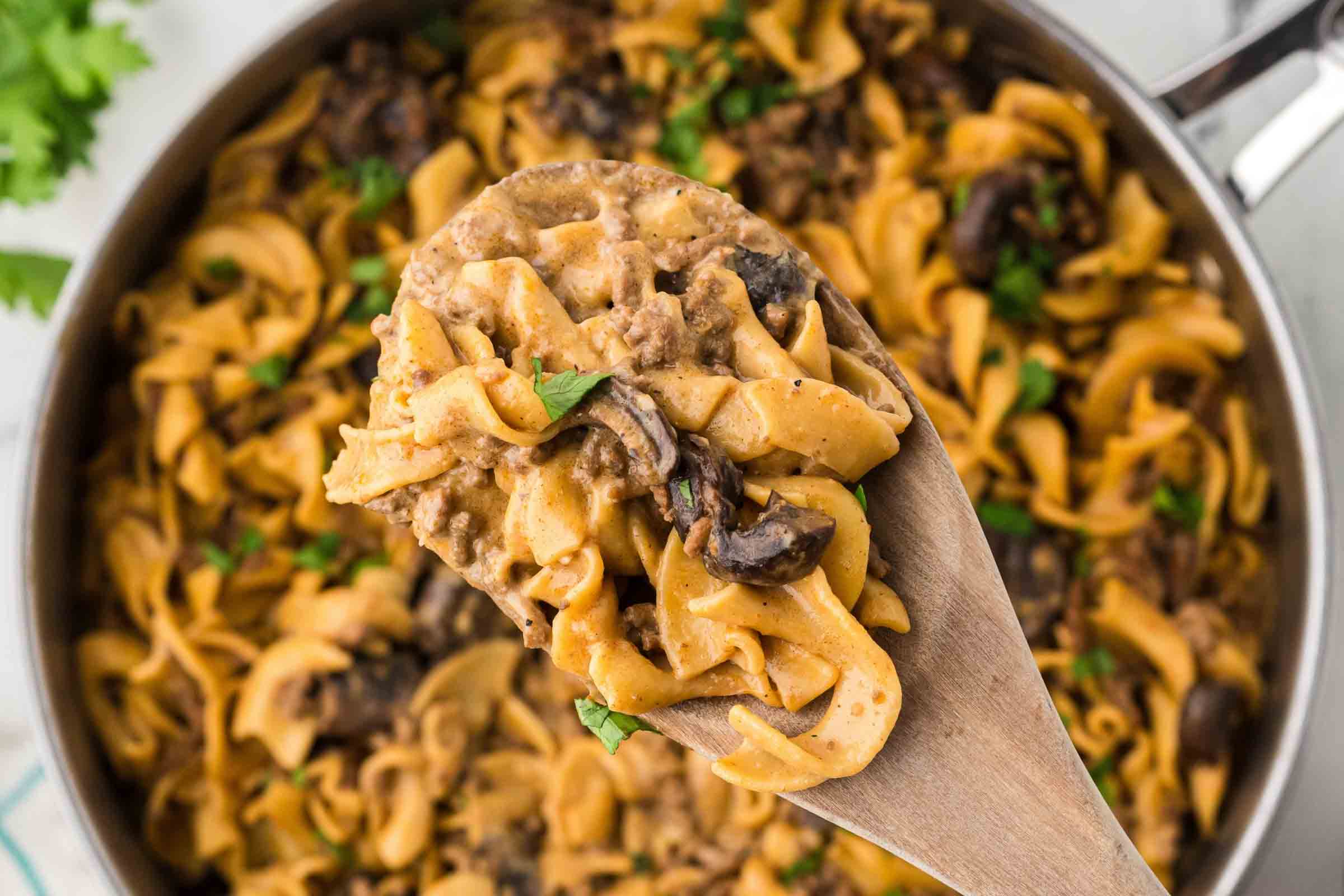 A pan with a finished recipe showing egg noodles, sliced mushrooms, and ground beef in a sauce garnished with chopped parsley with a serving lifted toward the camera on a wooden spoon.
