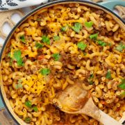 Skillet of cheesy elbow macaroni and ground beef in seasoned tomatoes garnished with cilantro with a wooden spoon.