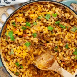 Skillet of cheesy elbow macaroni and ground beef in seasoned tomatoes garnished with cilantro with a wooden spoon.