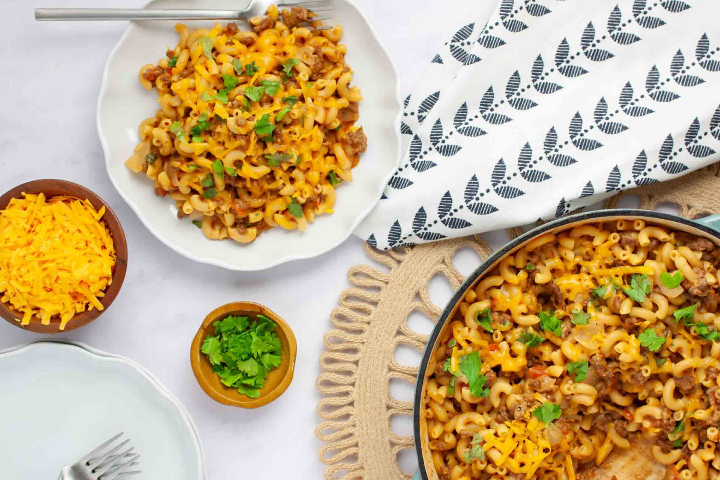 Plate of cheesy elbow macaroni and ground beef in seasoned tomatoes garnished with cilantro beside a pan of the same.