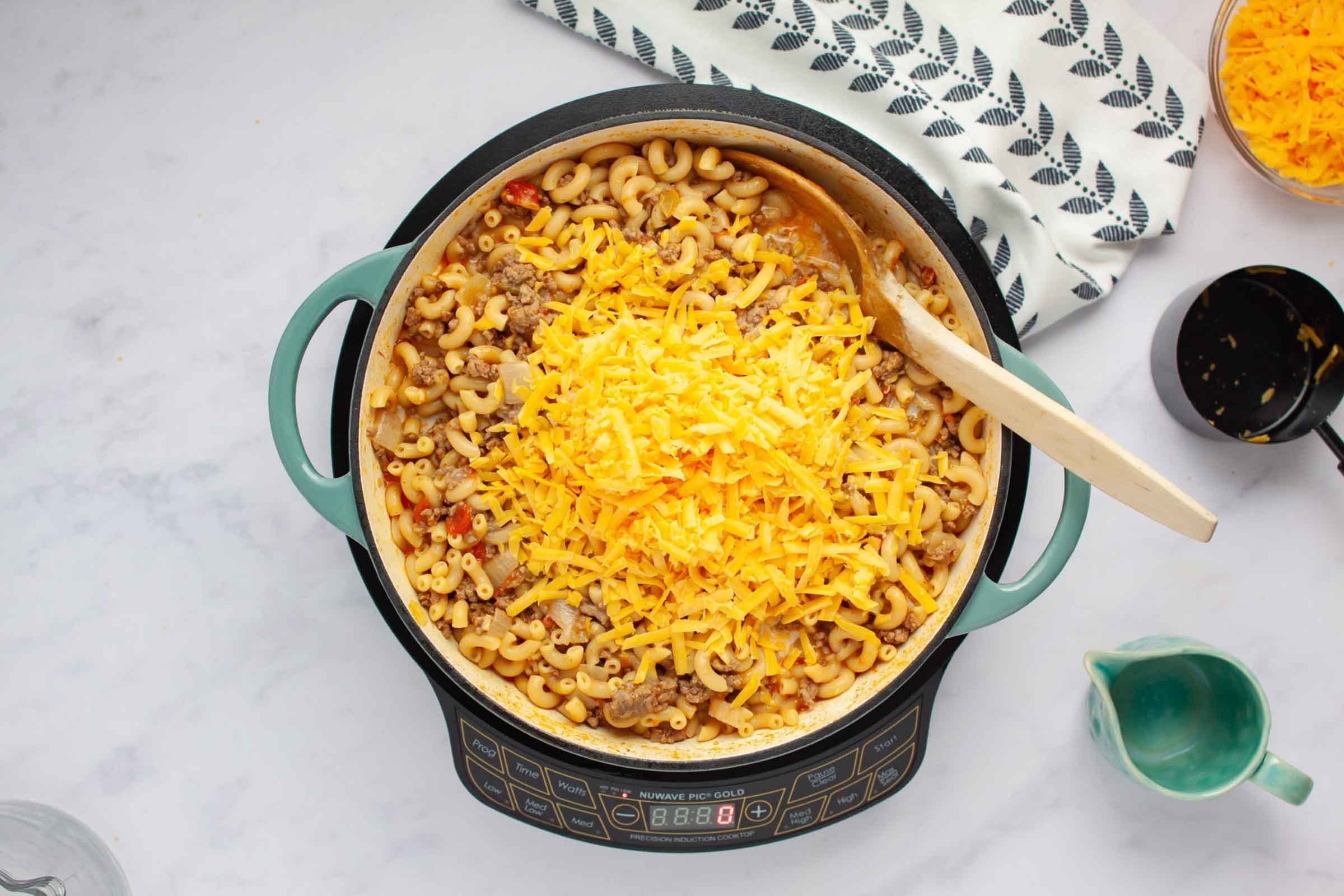 Skillet with ground beef and elbow macaroni in a seasoned tomato sauce with shredded cheddar cheese added but not stirred in.