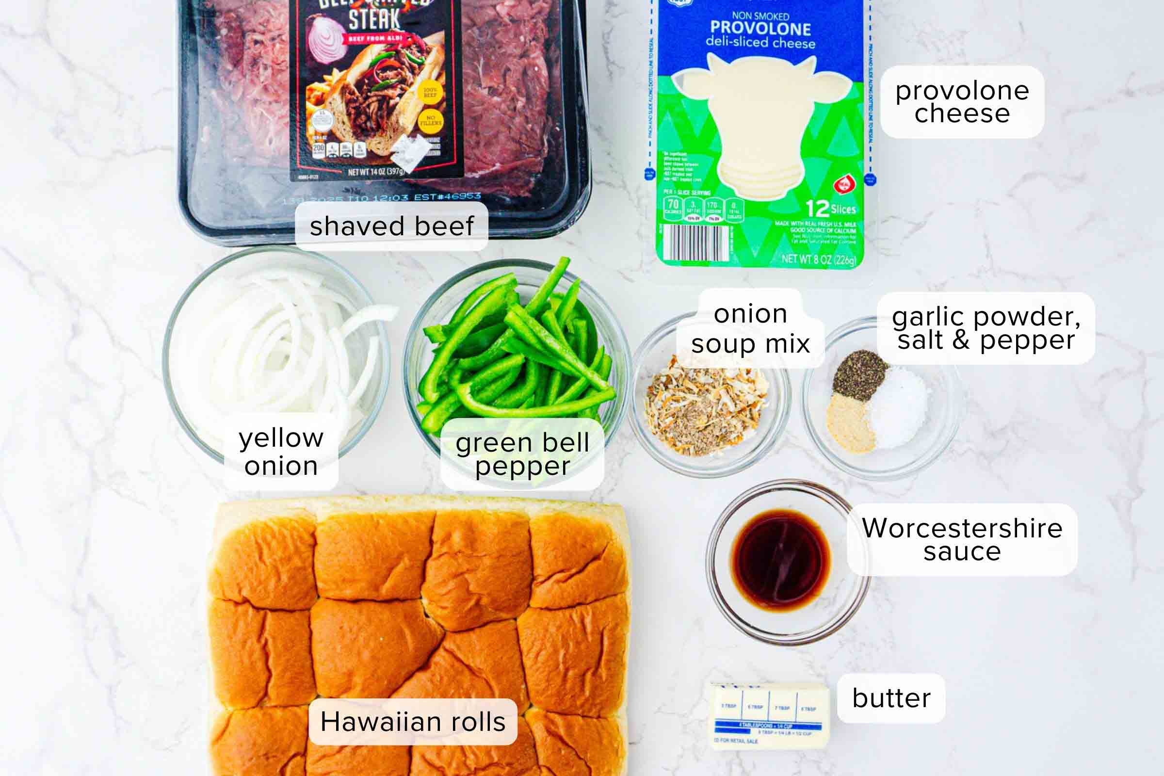 Labeled ingredients on a counter: shaved beef, provolone cheese slices, sliced yellow onion, sliced green bell pepper, onion soup mix, garlic powder, salt, pepper, Worcestershire sauce, butter, and Hawaiian rolls.
