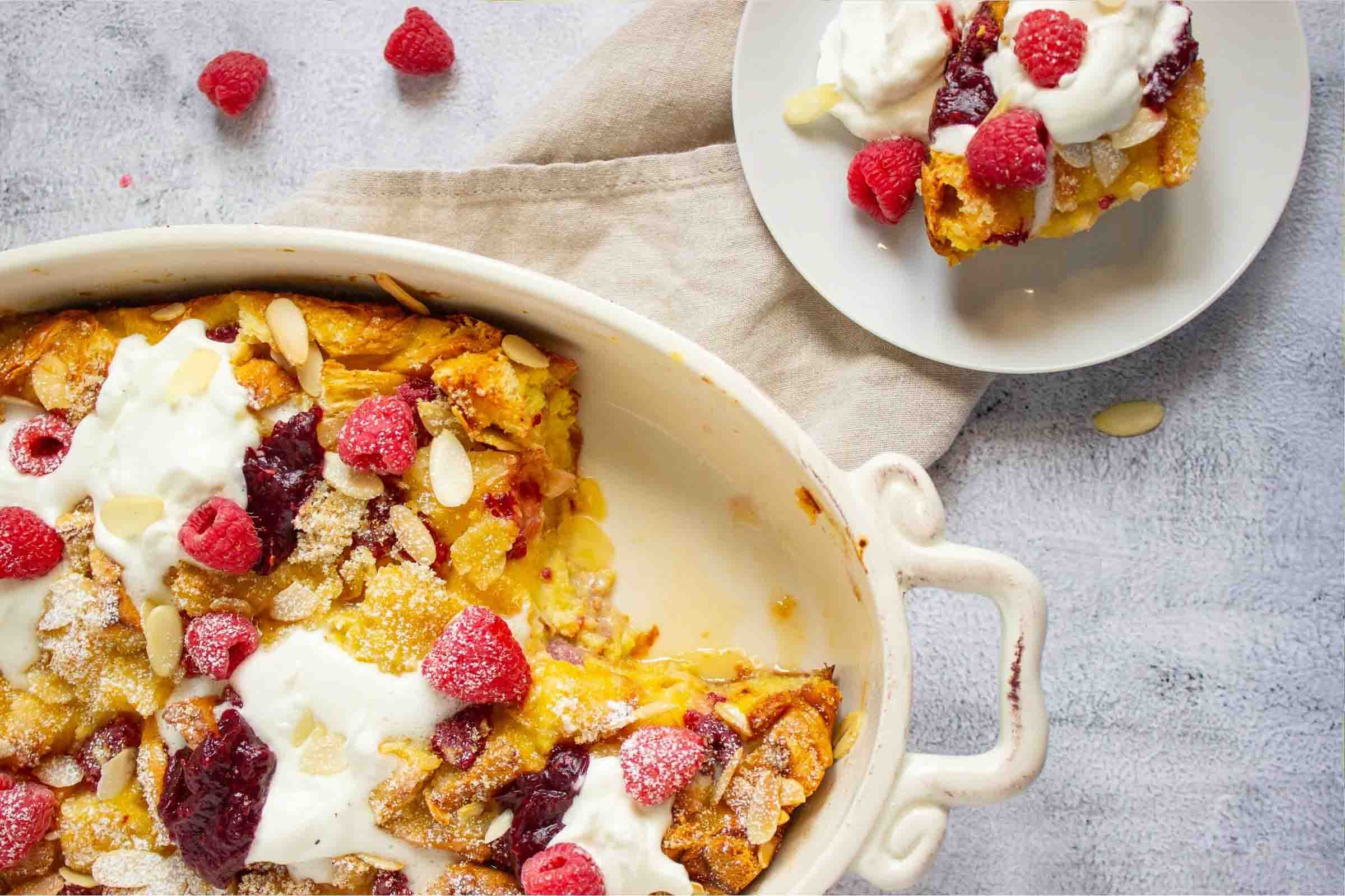 French toast bake with berries and almonds garnished with powdered sugar, whipped cream, and jam in a casserole dish with a serving on a plate.