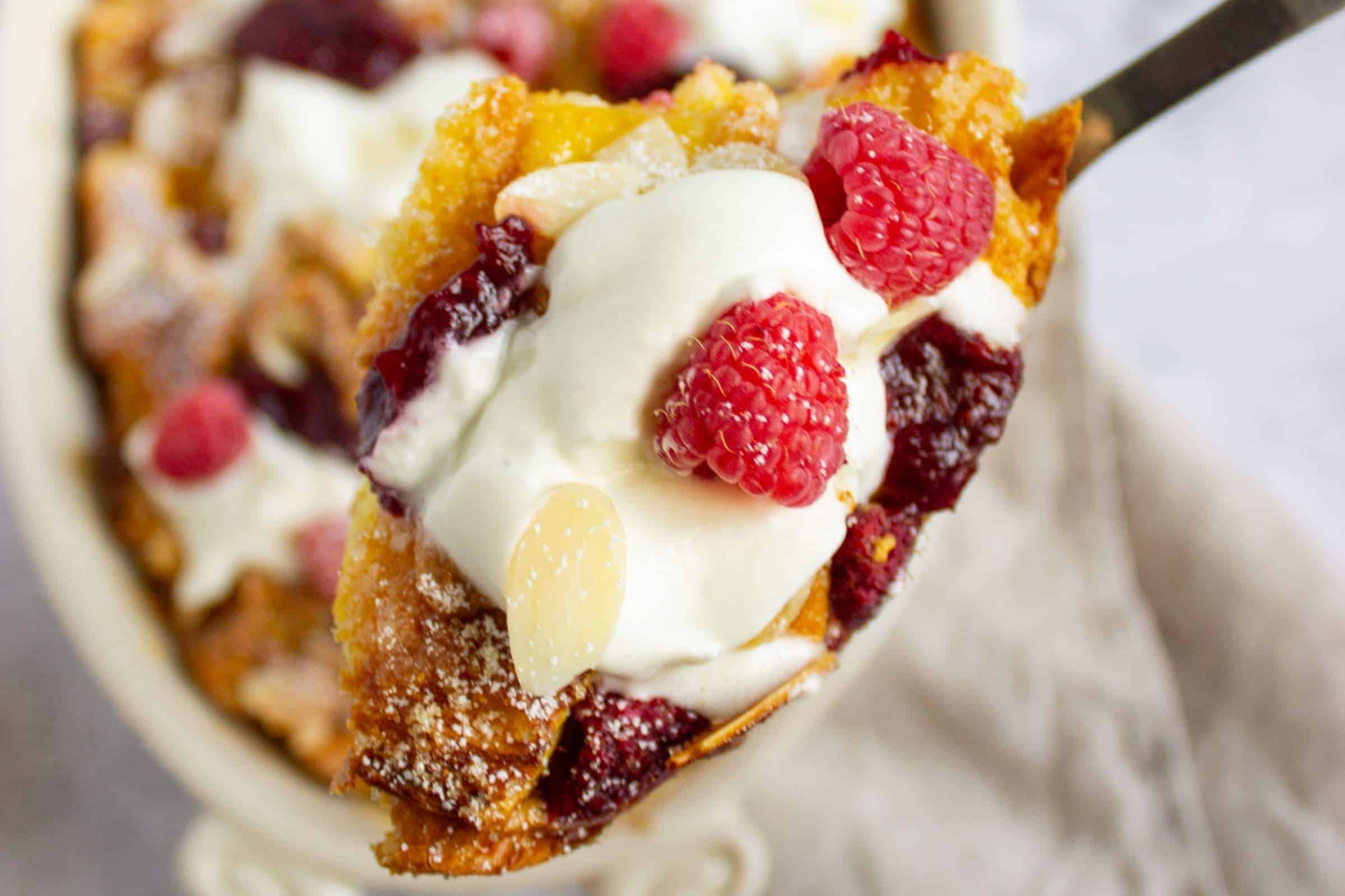 French toast bake with berries and almonds garnished with powdered sugar, whipped cream, and jam in a casserole dish with a serving held toward the camera.