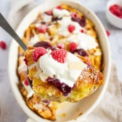 French toast bake with berries and almonds garnished with powdered sugar, whipped cream, and jam in a casserole dish with a serving held toward the camera.