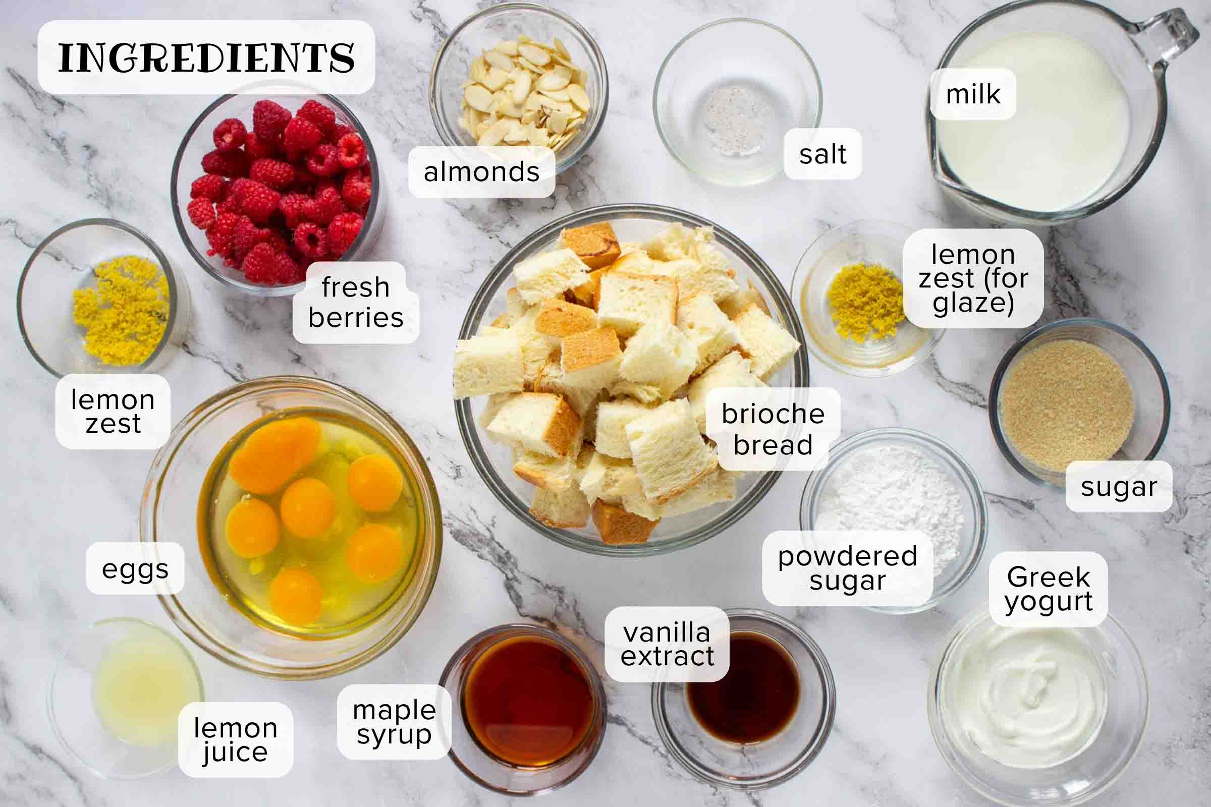 Ingredients on a counter: brioche bread, eggs, lemon juice, maple syrup, vanilla extract, powdered sugar, Greek yogurt, sugar, lemon zest, milk, salt, almonds, and berries.