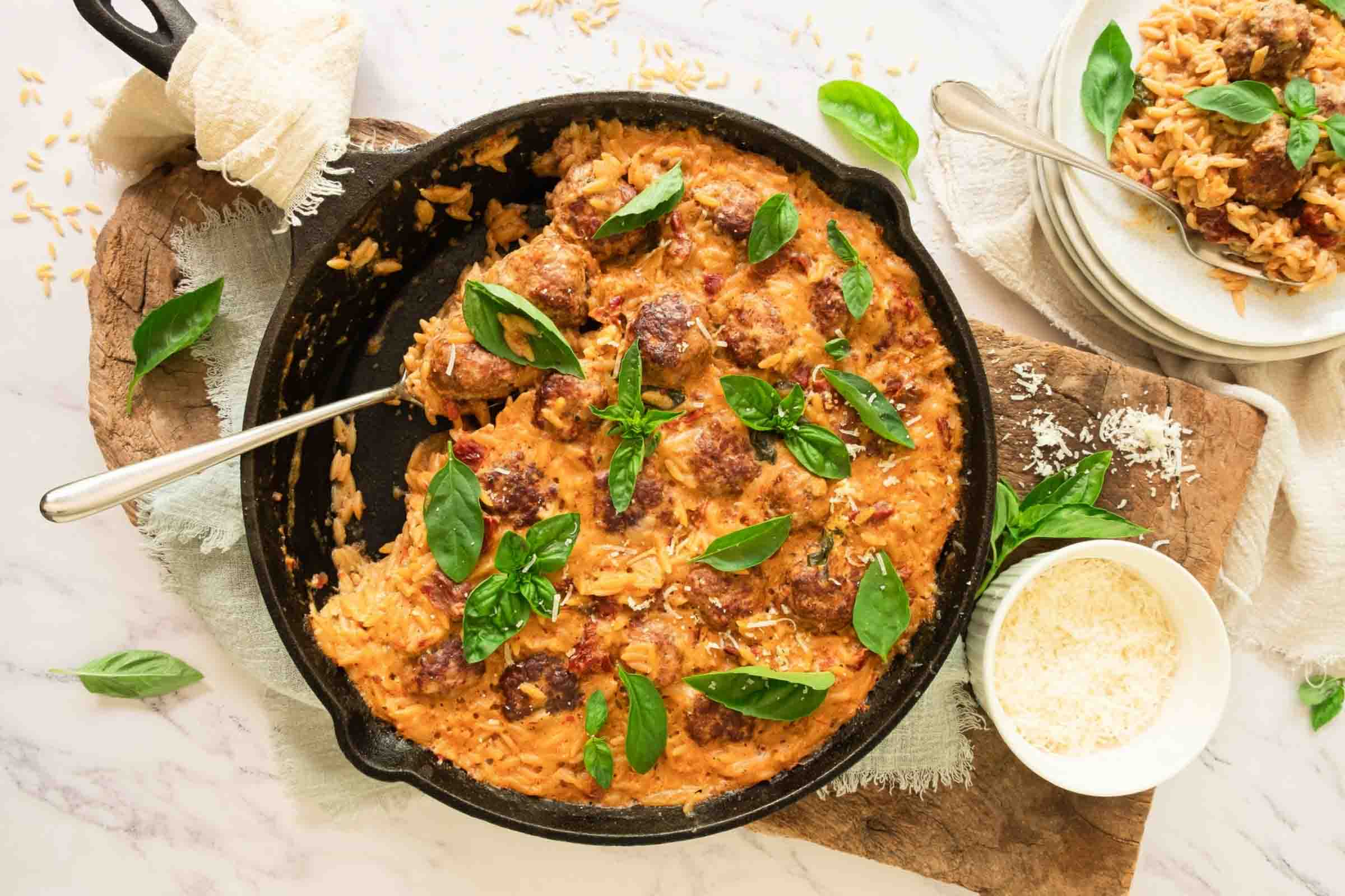A cast iron skillet of meatballs with orzo in a creamy sun-dried tomato sauce and garnished with fresh basil.