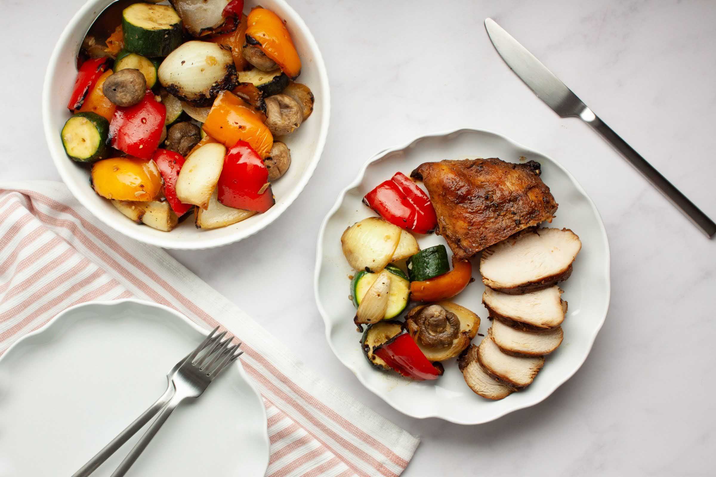 Sliced piece of grilled sesame chicken on a plate with grilled vegetables beside a bowl of grilled vegetables.